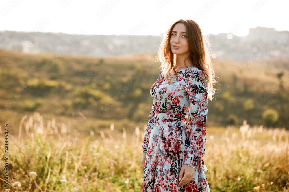 beautiful young woman in dress in floral print walking in the field at sunset. stylish romantic girl with long hair have a good time outdoors on city background. copy space beautiful young woman in dress in floral print walking in the field at sunset. stylish romantic girl with long hair have a good time outdoors on city background. copy space