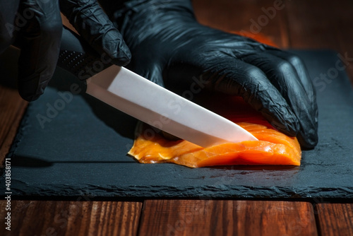 Chef use knife to cutting salmon fillet on black board at professional kitchen. Closeup chef hands in black gloves slicing red fish slice