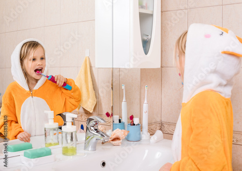 Five year old girl wearing orange pyjamas brushes her teeth with electric toothbrush in a bathroom photo