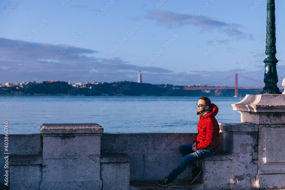 A man in sunglasses, a red jacket and jeans sits by the river on the embankments.