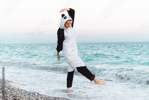 Emotional girl wears kigurumi laughing in front of the camera. Adorable young woman by the sea in panda pajamas dancing