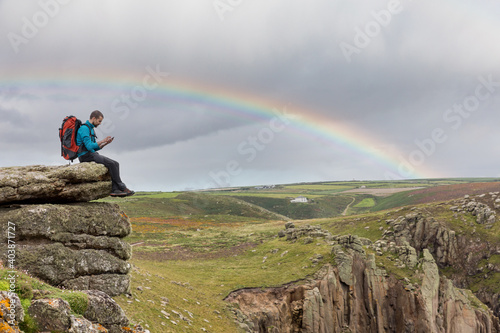 Fototapeta Man sitting on a rock cliff  and using his phone - Young man with backpack and h