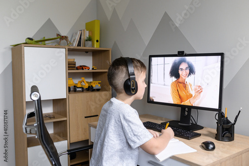 Above shoulder shot of a cute schoolkid watching online video classes sitting on self isolation at home, a smiling female teacher on the screen. E-learning, distance education