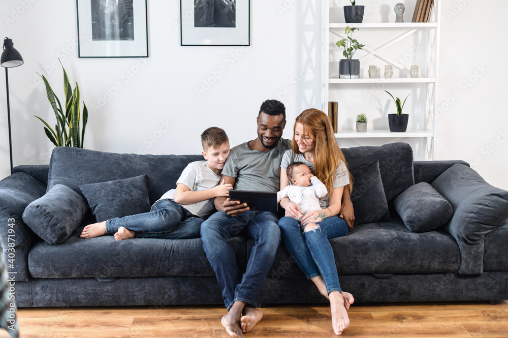 A multiracial family relaxing with a digital tablet sitting on the comfortable sofa, watching video, playing game on the touchscreen, have fun together. Spouses, baby girl and school-age son A multiracial family relaxing with a digital tablet sitting on the comfortable sofa, watching video, playing game on the touchscreen, have fun together. Spouses, baby girl and school-age son
