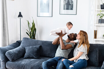 A multiracial family on the comfortable couch at home. A father throwing his baby daughter up, a mother sits near and smiles