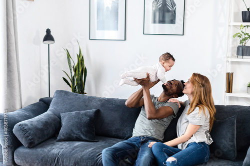 A multiracial family on the comfortable couch at home. A father throwing his baby daughter up, a mother sits near and smiles