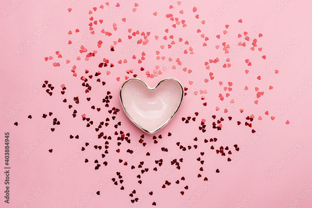 Pink ceramic heart shaped bowl with splash of red heart shaped confetti over pink background. Top view, Valentine's Day concept