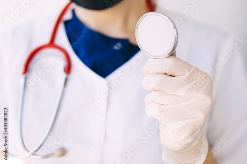 young doctor in a medical coat, gloves and mask holds a red stethoscope, listens to a patient with coronavirus