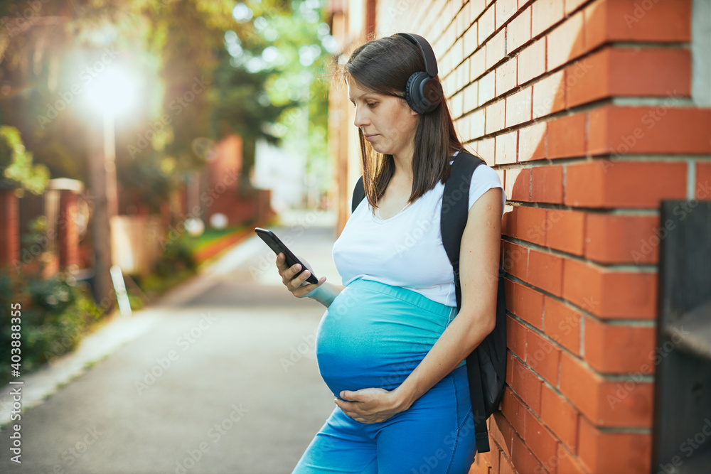 A pregnant sportswoman standing against the wall outdoors with headphones on ears and using smart phone.