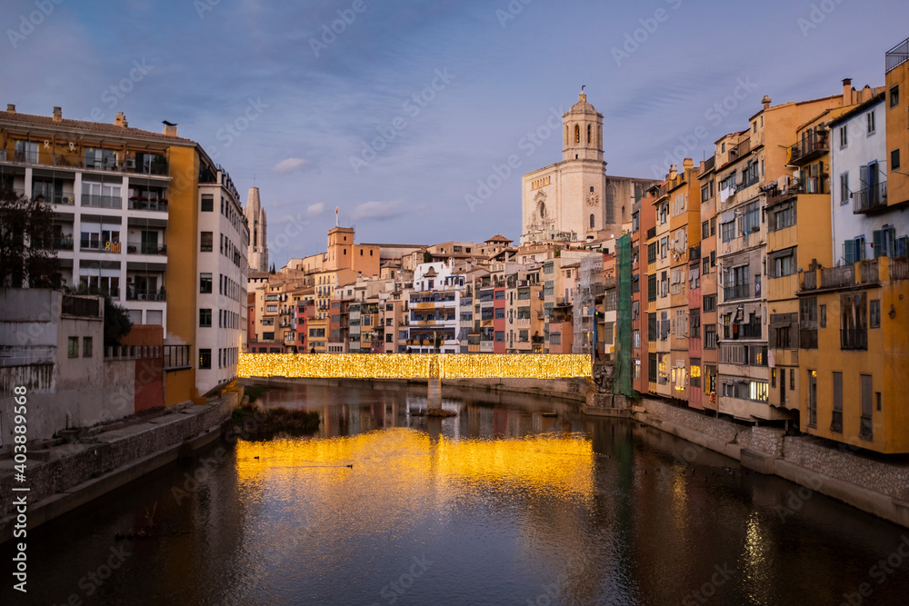 Girona's urban cityscape skyline at dusk with famous gothic cathedral landmark and river houses reflected on a quiet river from red iron bridge