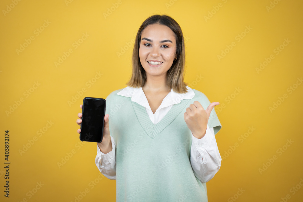 Pretty young woman standing over yellow background holding smartphone showing blank screen smiling happy pointing with hand and finger to the side Pretty young woman standing over yellow background holding smartphone showing blank screen smiling happy pointing with hand and finger to the side