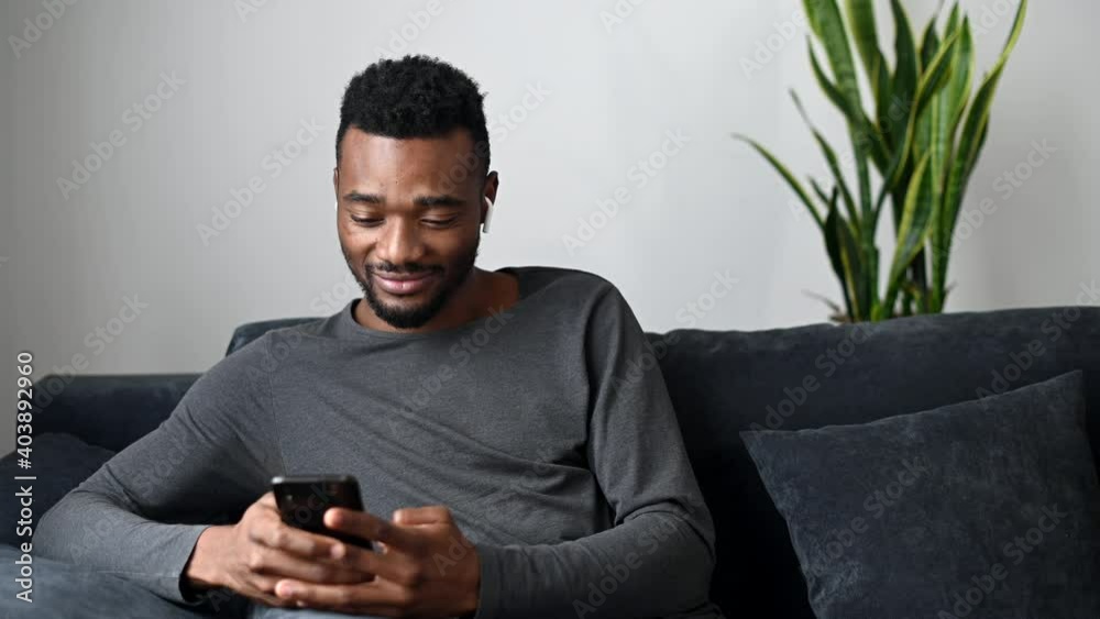 An African-American guy using smartphone for messaging, scrolling news, reading feeds in social media sitting on the couch at living room. Front view of a smiling multiracial millennial with a phone