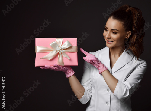 Young pretty brunette woman doctor gynecologist reproductologist in pink latex gloves and white uniform standing and holding holiday present box over dark background. Gynecology concept photo