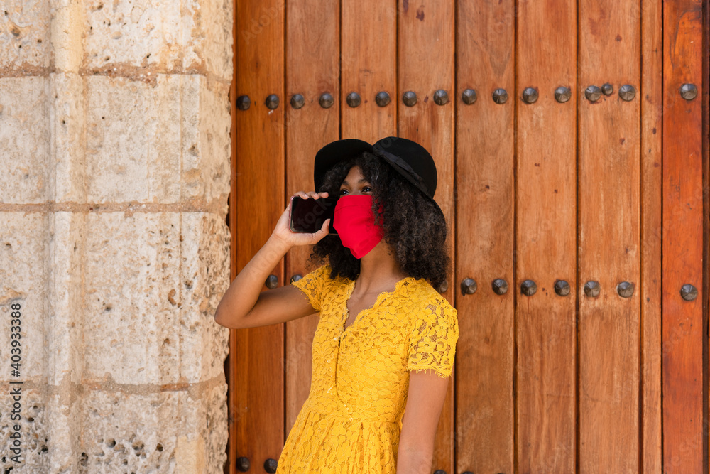 Obraz premium young black woman with curly hair, with a red mask, yellow dress and black hat, talking with her smart phone, in front of an old door