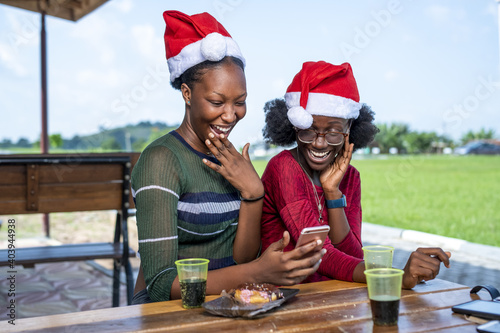 Positive Black friends in Santa hats having a video call while sitting outdoors at a picnic table photo