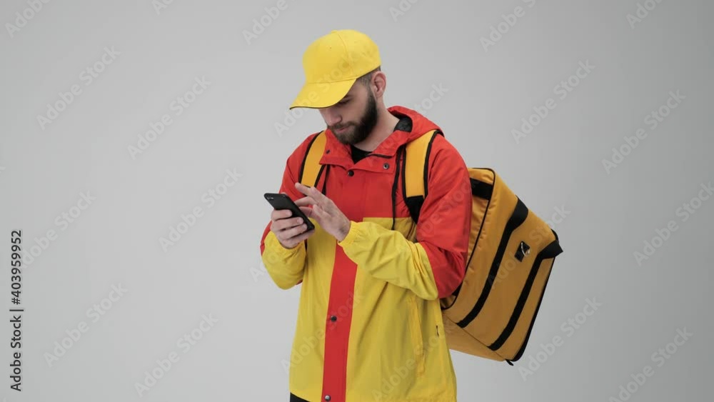 Attractive courier in red yellow uniform with cap and backpack sending message on his smartphone zooming screen nodding head studio white background. Online order express deliver home food Attractive courier in red yellow uniform with cap and backpack sending message on his smartphone zooming screen nodding head studio white background. Online order express deliver home food