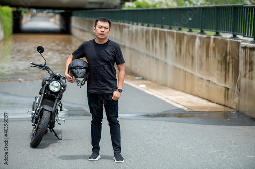 Confident asian man with helmet in hands standing next to his motorcycle 