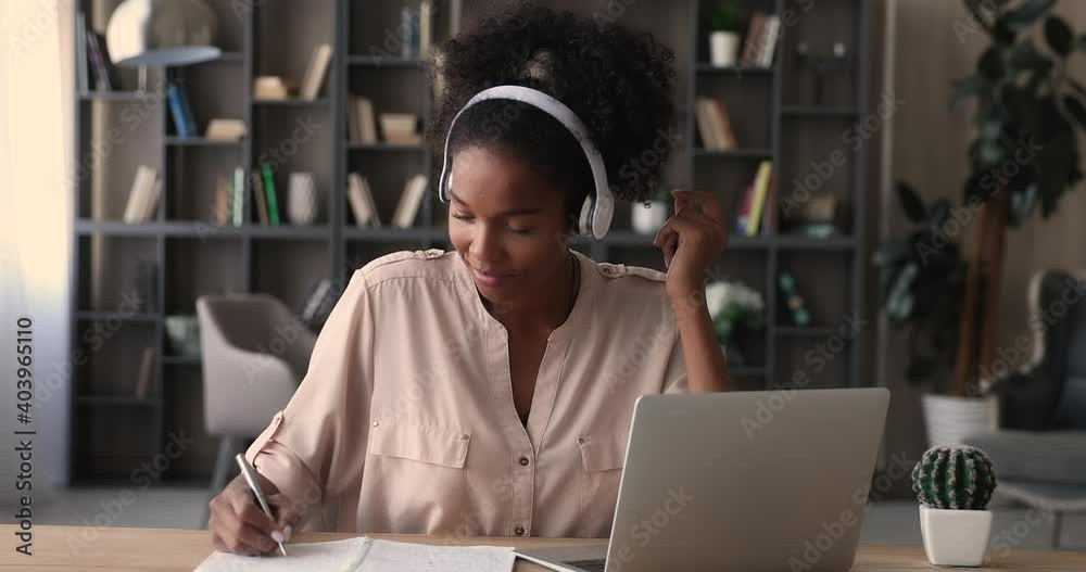 Attractive mixed race woman wear cordless headphones listen audio course doing task, takes useful information on website, making notes, studying on-line using internet resources for students concept