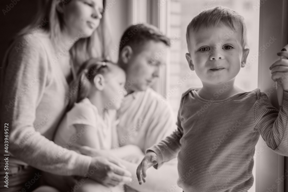 cute caucasian little boy standing on window sill. His parents and sister on background looking out the window. Home isolation during pandemia. Black and white image with selectie focus cute caucasian little boy standing on window sill. His parents and sister on background looking out the window. Home isolation during pandemia. Black and white image with selectie focus