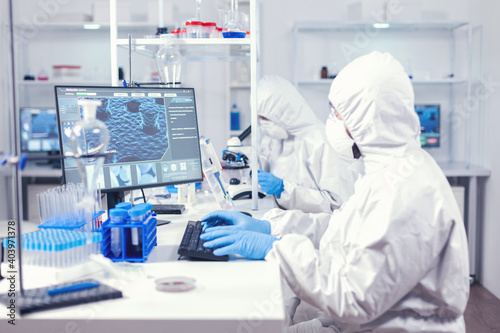 Lab engineer typing on pc conducting experiment in laboratory dressed in ppe suit. Medical engineer using computer during global pandemic with coronavirus dressed in coverall.