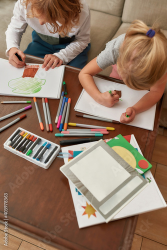 High angle view of focused little kids, boy and girl drawing on paper using marker pen, sitting together on a couch at home