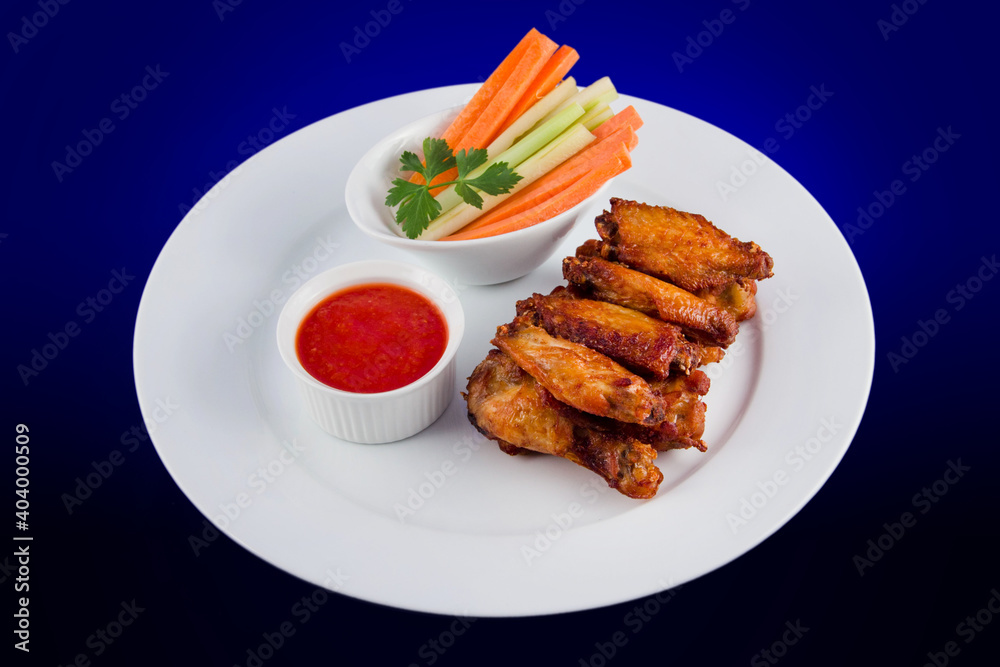 Roasted chicken wings with sticks carrots and celery and dipping sauce on white plate on blue background Roasted chicken wings with sticks carrots and celery and dipping sauce on white plate on blue background