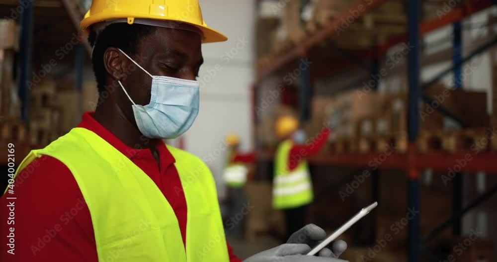 African worker using tablet inside warehouse while wearing protective face mask for coronavirus prevention African worker using tablet inside warehouse while wearing protective face mask for coronavirus prevention