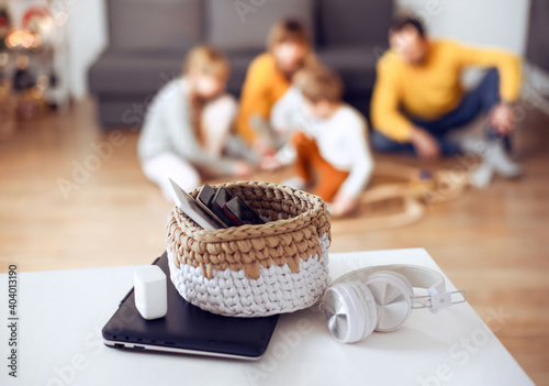 Mobile phones, laptops and digital tablets in a basket on table at home with playing family at back. photo
