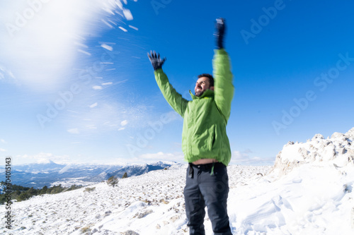 Happy brunette handsome man playing in snow wearing green hiking jacket smiling on snowy mountain