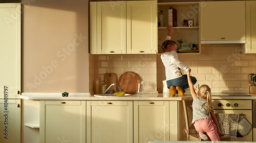 Schilderij op canvas Rear view of two naughty kids, brother and sister trying to find something sweet in the kitchen cabinet