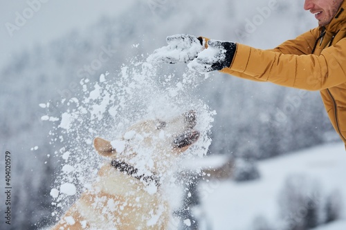 Young man with happy dog in winter. Pet owner with his labrador retriever playing in snow in beautiful nature. 