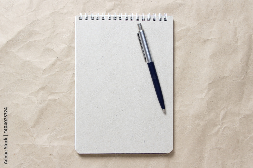 Gray notepad with white coiled spring and pen on a background of beige crumpled craft paper. With empty space for text and design Gray notepad with white coiled spring and pen on a background of beige crumpled craft paper. With empty space for text and design