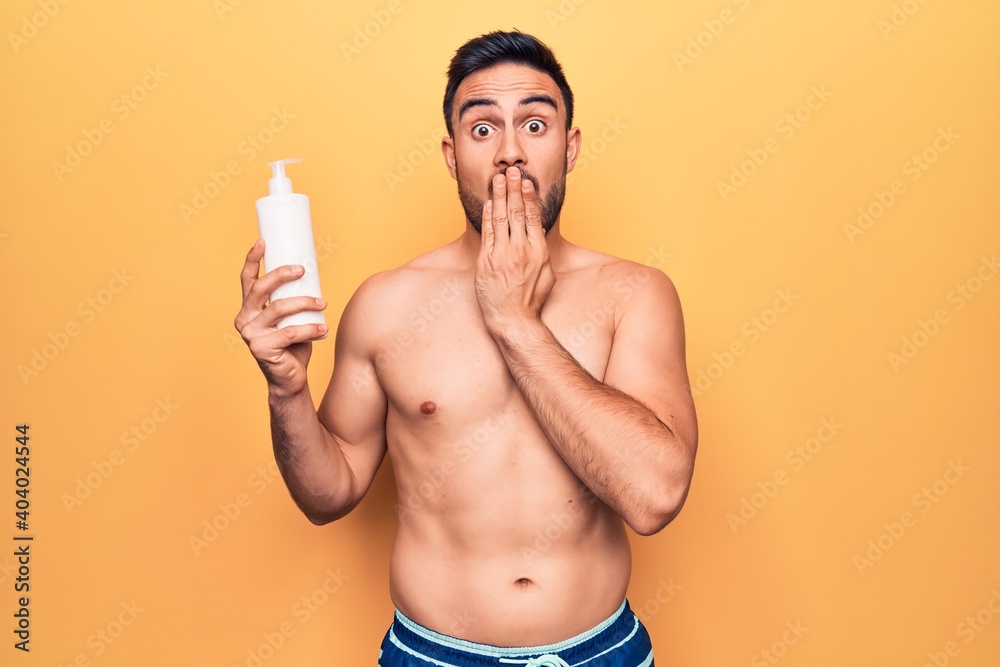 Fototapeta premium Handsome man with beard on vacation wearing swimwear holding bottle of sunscreen lotion covering mouth with hand, shocked and afraid for mistake. Surprised expression