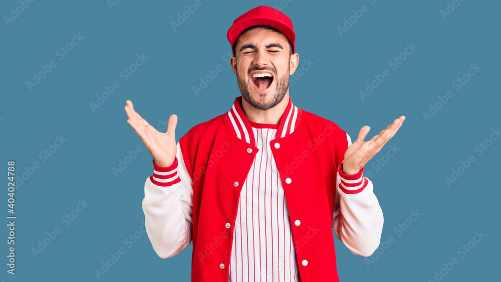 Young handsome man wearing baseball jacket and cap celebrating mad and crazy for success with arms raised and closed eyes screaming excited. winner concept Young handsome man wearing baseball jacket and cap celebrating mad and crazy for success with arms raised and closed eyes screaming excited. winner concept