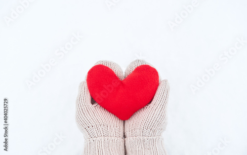 Creative greeting card for Valentines Day. Hold red soft heart toy made of fabric in beige mittens against background of white freshly fallen snow. Empty space for your greeting text.