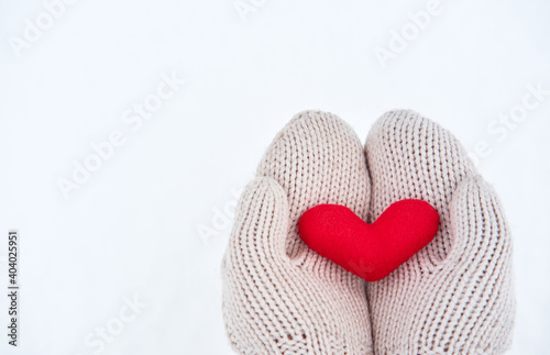 Creative greeting card for Valentines Day. Holding red little soft fabric toy heart in beige mittens against background of white freshly fallen snow. Empty space for your greeting text.