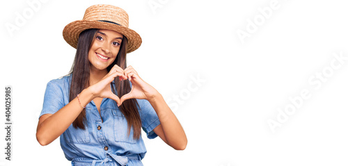 Young hispanic woman wearing summer hat smiling in love showing heart symbol and shape with hands. romantic concept.