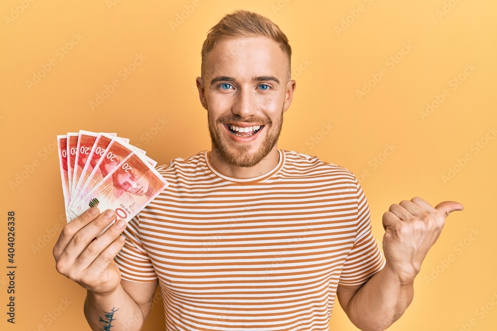 Young caucasian man holding 20 israel shekels banknotes pointing thumb up to the side smiling happy with open mouth Young caucasian man holding 20 israel shekels banknotes pointing thumb up to the side smiling happy with open mouth
