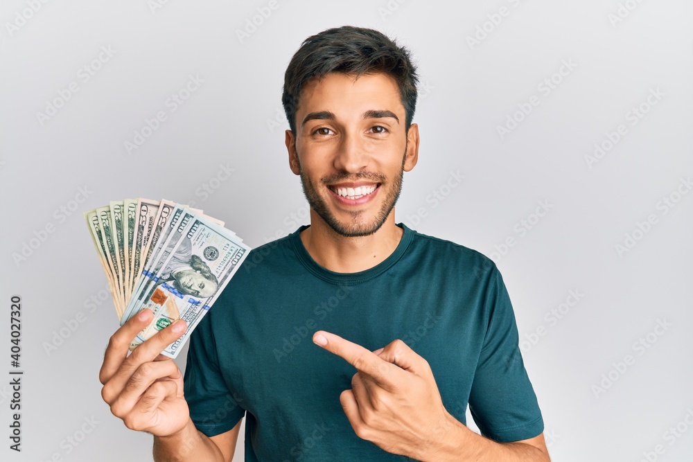 Young handsome man holding dollars smiling happy pointing with hand and finger Young handsome man holding dollars smiling happy pointing with hand and finger