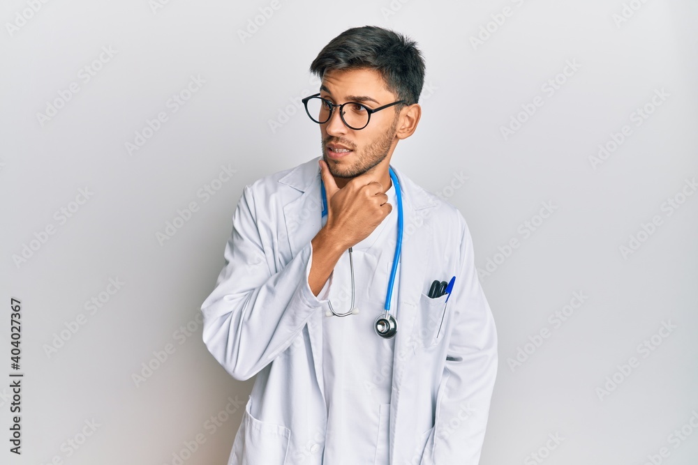 Young handsome man wearing doctor uniform and stethoscope with hand on chin thinking about question, pensive expression. smiling with thoughtful face. doubt concept. Young handsome man wearing doctor uniform and stethoscope with hand on chin thinking about question, pensive expression. smiling with thoughtful face. doubt concept.