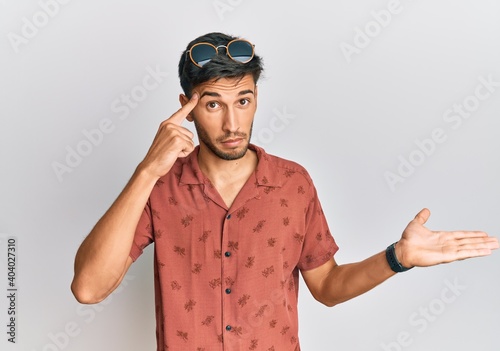 Young handsome man wearing casual summer clothes confused and annoyed with open palm showing copy space and pointing finger to forehead. think about it.