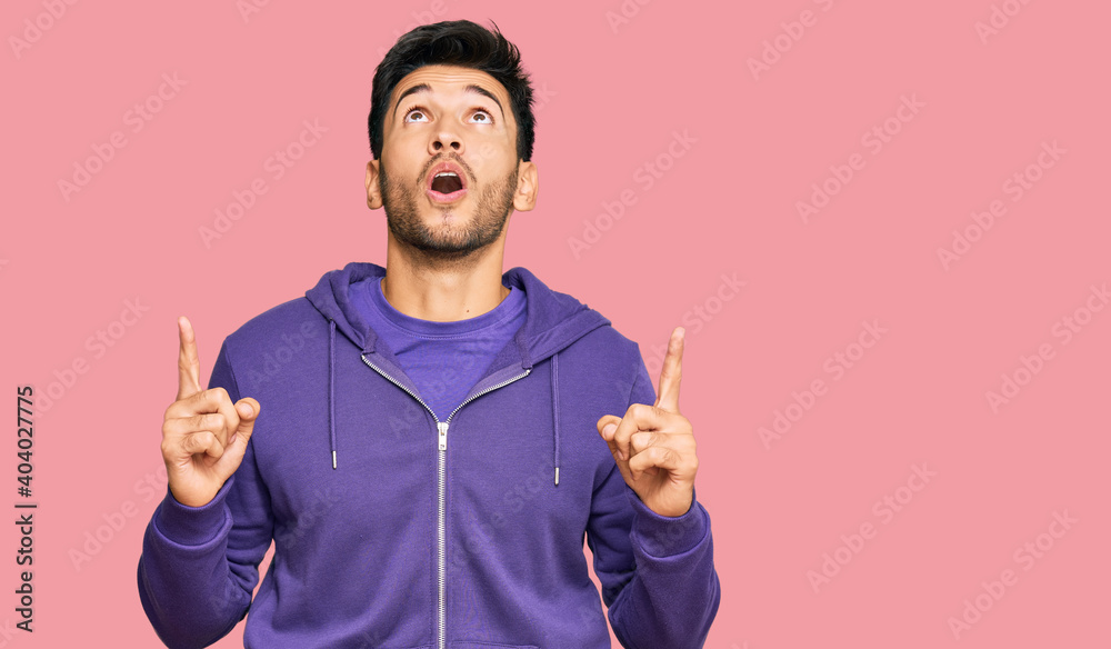 Young handsome man wearing casual sweatshirt amazed and surprised looking up and pointing with fingers and raised arms. Young handsome man wearing casual sweatshirt amazed and surprised looking up and pointing with fingers and raised arms.