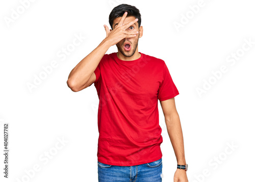 Young handsome man wearing casual red tshirt peeking in shock covering face and eyes with hand, looking through fingers with embarrassed expression.