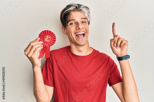 Young hispanic man holding second place badge smiling with an idea or question pointing finger with happy face, number one