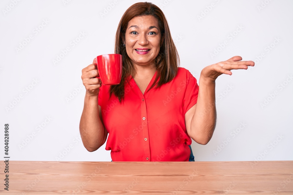Middle age brunette woman drinking a cup of coffee sitting on the table celebrating victory with happy smile and winner expression with raised hands Middle age brunette woman drinking a cup of coffee sitting on the table celebrating victory with happy smile and winner expression with raised hands