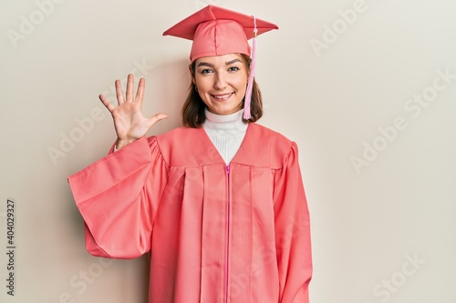 Young caucasian woman wearing graduation cap and ceremony robe showing and pointing up with fingers number five while smiling confident and happy.