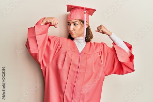 Young caucasian woman wearing graduation cap and ceremony robe showing arms muscles smiling proud. fitness concept.