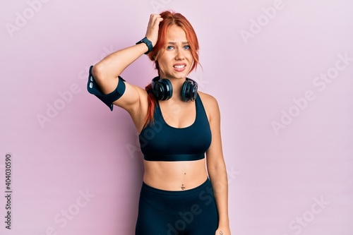 Young beautiful redhead woman wearing gym clothes and using headphones confuse and wonder about question. uncertain with doubt, thinking with hand on head. pensive concept.