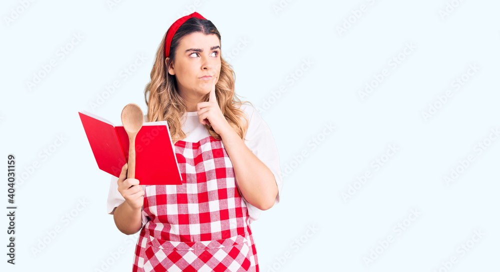 Young caucasian woman wearing apron holding recipe book and spoon serious face thinking about question with hand on chin, thoughtful about confusing idea Young caucasian woman wearing apron holding recipe book and spoon serious face thinking about question with hand on chin, thoughtful about confusing idea