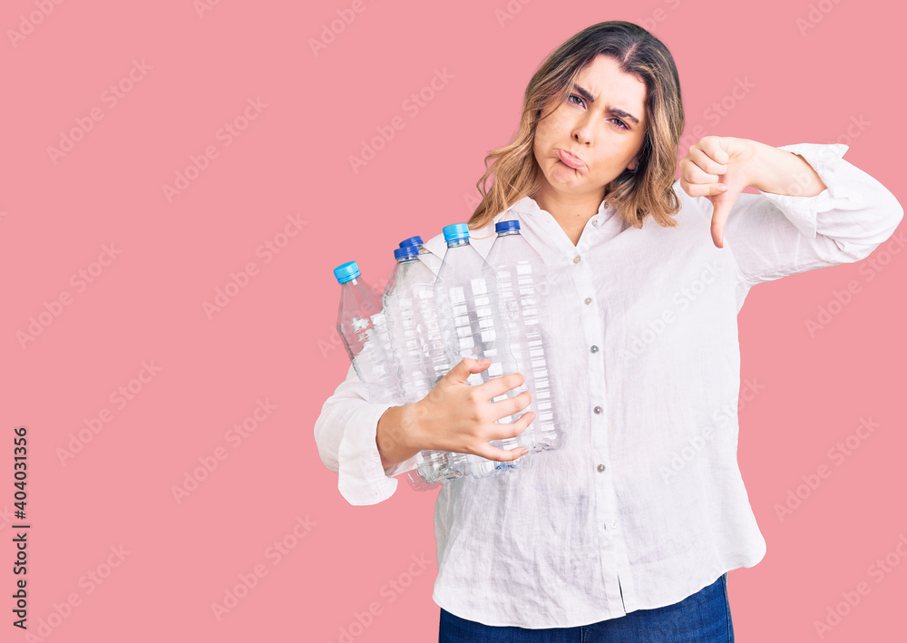 Young caucasian woman holding recycling plastic bottles with angry face, negative sign showing dislike with thumbs down, rejection concept Young caucasian woman holding recycling plastic bottles with angry face, negative sign showing dislike with thumbs down, rejection concept